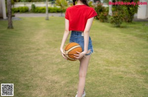 A woman leaning against a stone pillar in a red shirt and denim shorts.